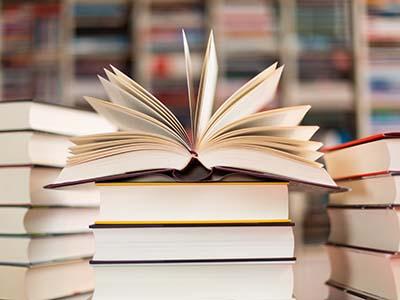 A stacxk of books on a table with the top book spread open. No covers can be seen. Bookshelves are in the back in the distant.