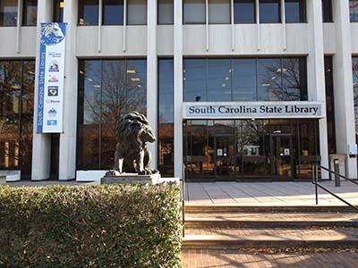 Exterior photo of the South Carolina State Library showing one of our lion statues out front, with the main door and sign above.