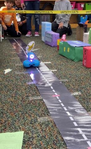 Students watch as a small robot progresses down a path lined with buildiings made from construction paper. 