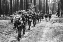 A black and white photo shows soldiers walking through the woods with their gear. 